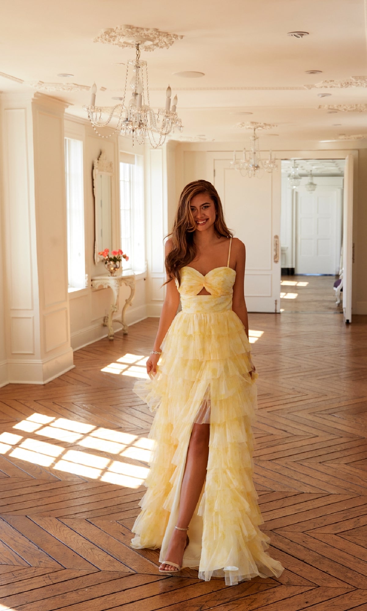 A woman wearing the Juliet JT2508A Long Yellow Ruffled Prom Dress with a tiered tulle A-line skirt and thin straps stands in a bright, elegant room with wooden floors and chandeliers.